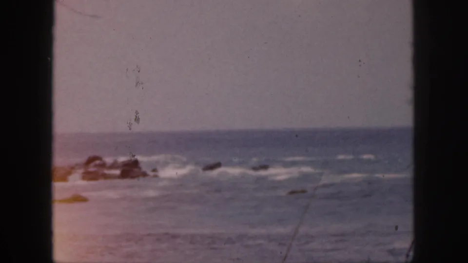1959 Hawaii shoreline view with rough waves around offshore rocks, soft focus, and dark 8mm frame borders.