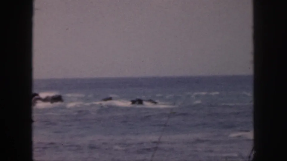 Faded 8mm Hawaii coastal frame showing a low horizon, choppy water, and surf around dark rocks.