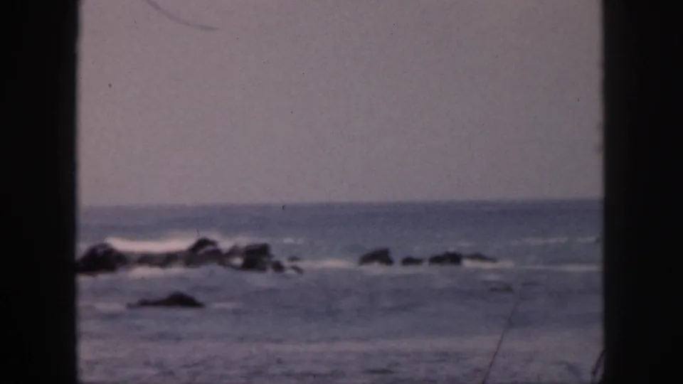 1959 Hawaii home-movie frame with white surf striking dark shoreline rocks under a hazy pale sky.