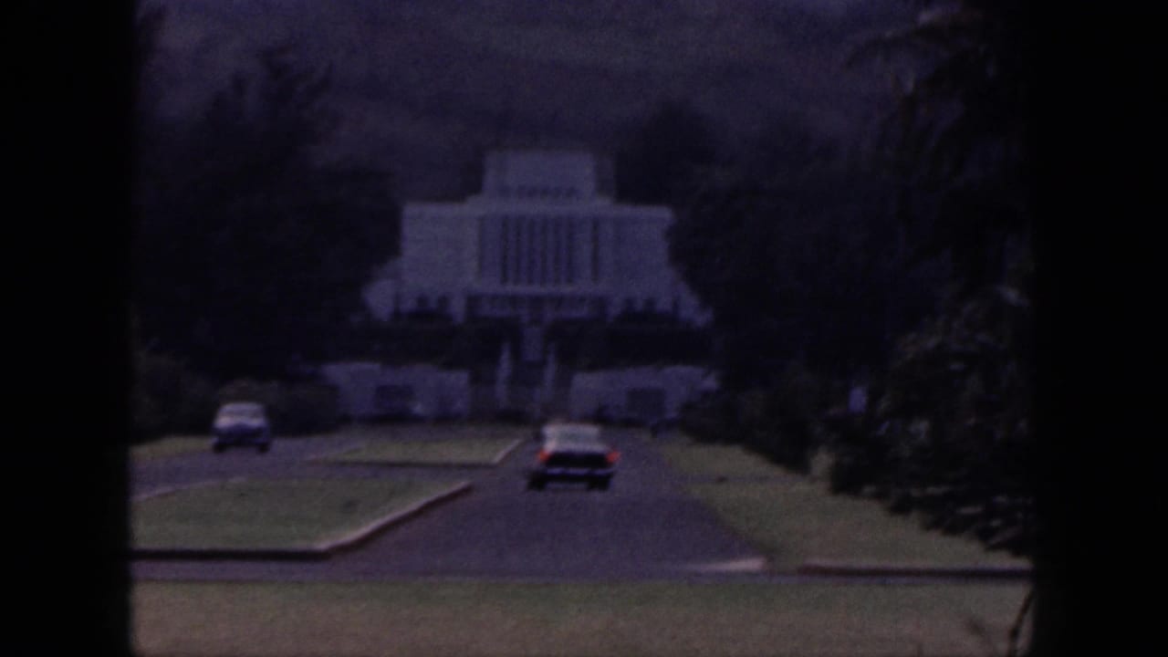1959 Hawaii 8mm frame showing classic cars approaching a white temple-like building set among lawns, trees, and hills.