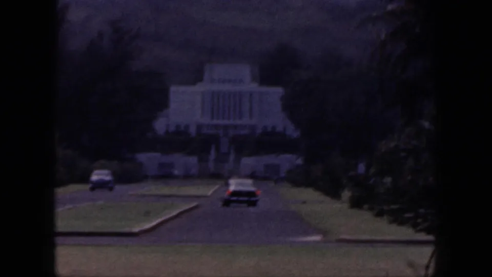 1959 Hawaii 8mm frame showing classic cars approaching a white temple-like building set among lawns, trees, and hills.
