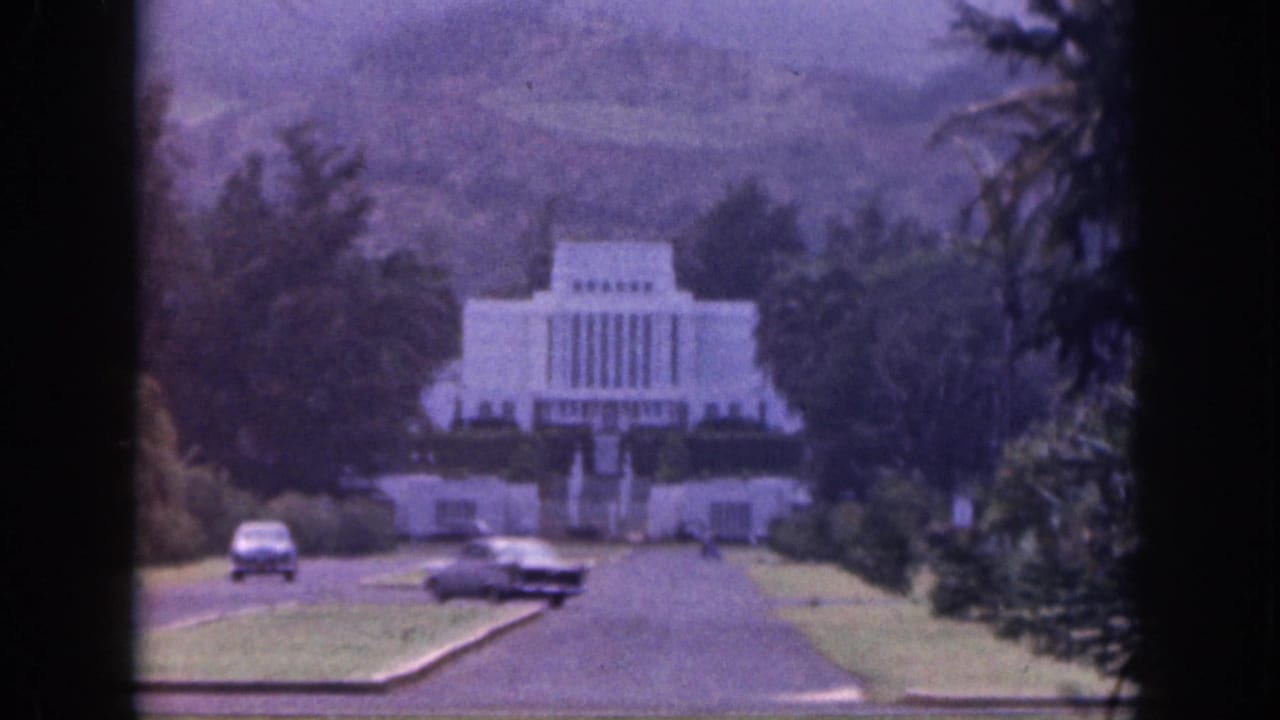 1959 Hawaii frame with two classic cars on a paved approach road below a white temple-like structure and wooded hillside.