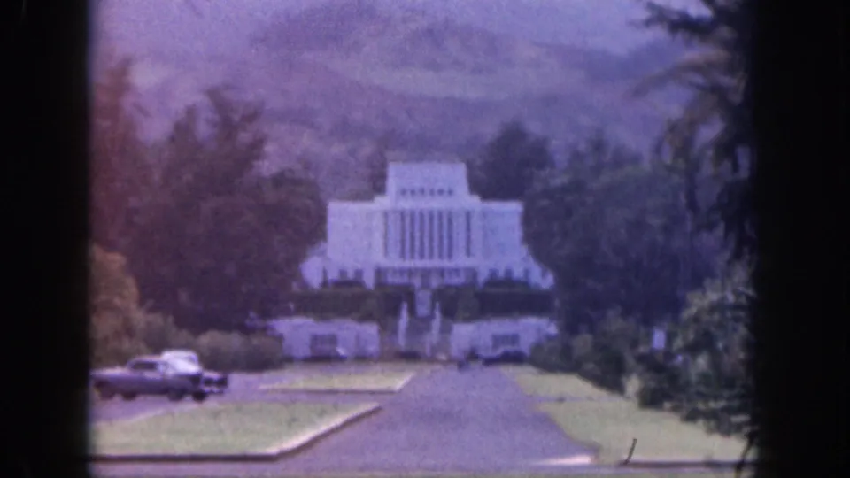 1959 Hawaii 8mm frame showing a classic car turning near lawns as a white temple-like building rises in the background.