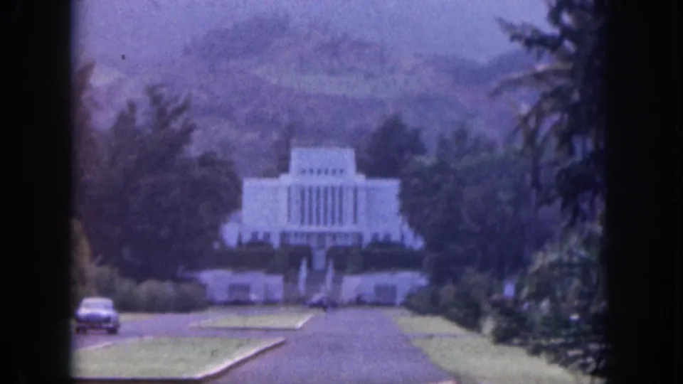 1959 Hawaii home-movie frame of a white temple-like building framed by greenery, with a classic car near the road.