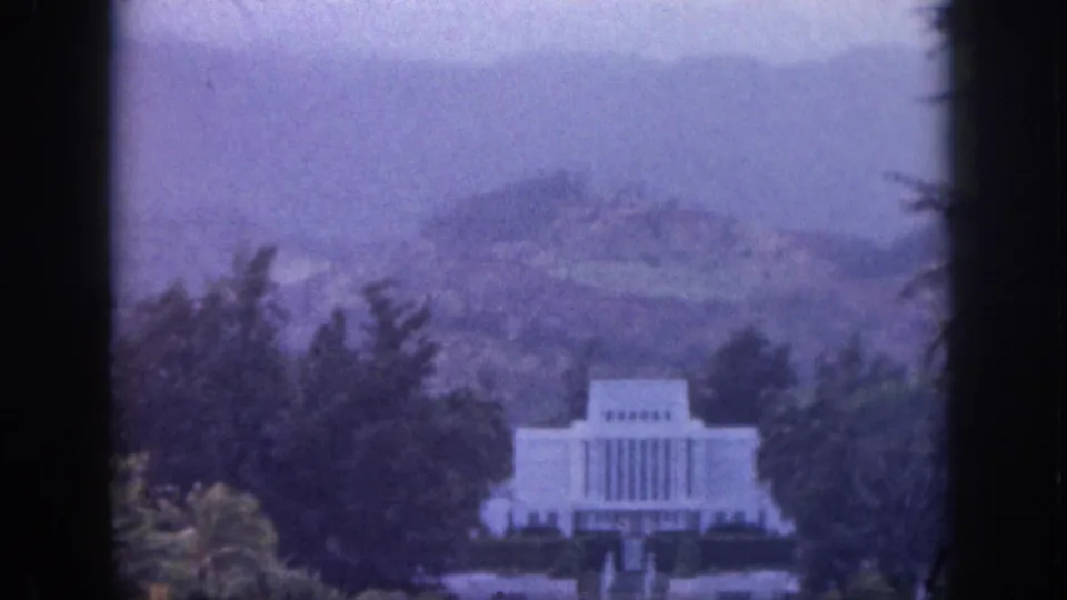 A soft, purple-blue 8mm frame from 1959 Hawaii shows a white temple-like building backed by trees and hazy hills.