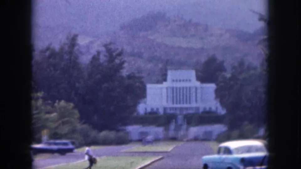 1959 Hawaii home-movie frame with a distant white landmark, classic cars, landscaped grounds, and one small pedestrian crossing the foreground.