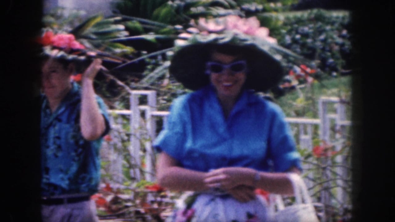 In 1959 Hawaii footage, two women in blue tops stand by a white fence wearing wide hats decorated with flowers and leaves.