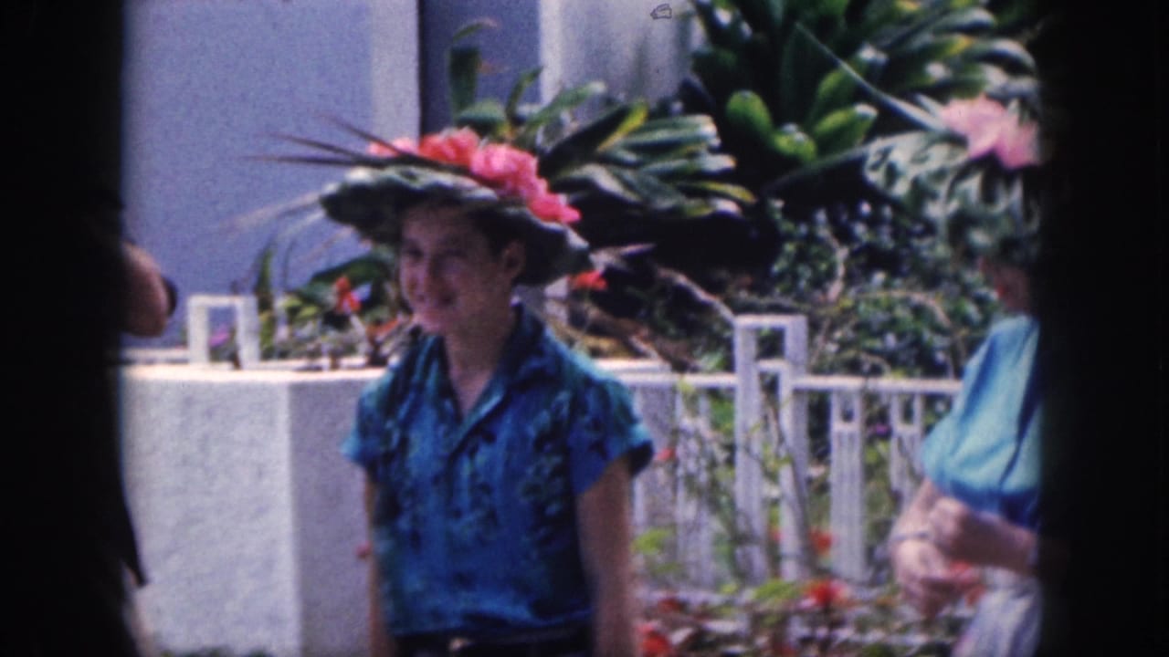 A woman in a blue patterned shirt stands near a white fence wearing a floral hat.