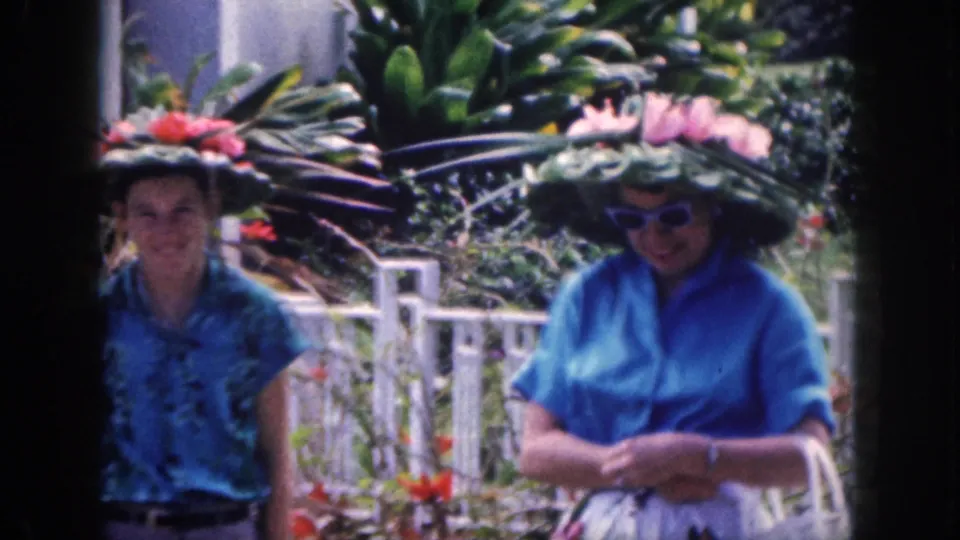 Two women in blue tops appear in 1959 Hawaii home-movie footage, wearing large floral hats among tropical plants and a white fence.