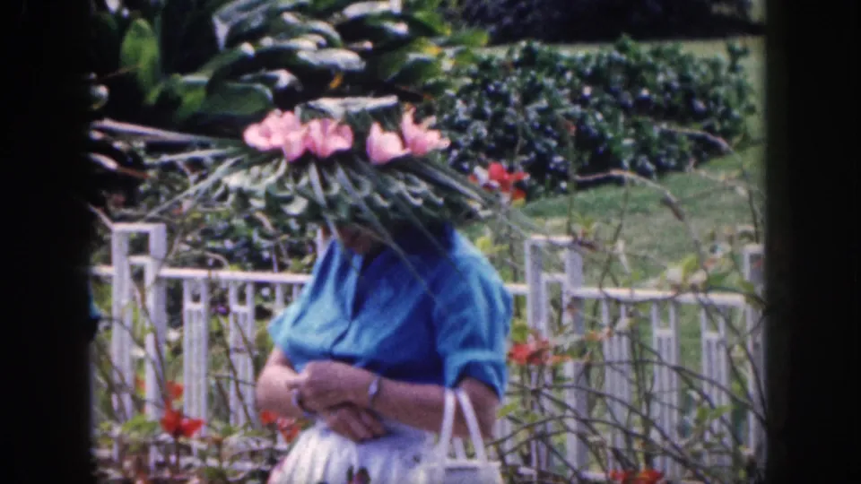 A woman in a blue blouse and sunglasses stands in the Hawaii garden with a white handbag and a wide hat covered in flowers and leaves.