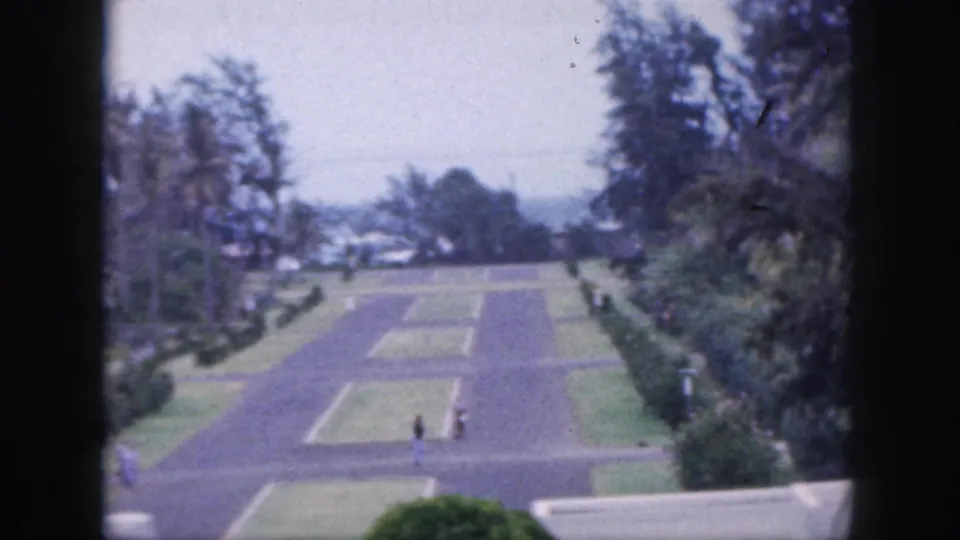 1959 Hawaii home-movie frame showing an elevated view over a formal garden path, rectangular lawns, trees, and small distant visitors.