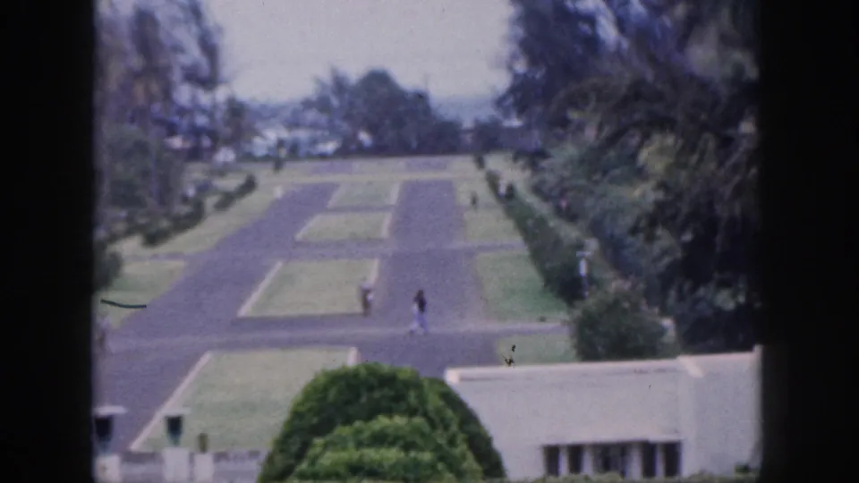1959 Hawaii home-movie frame with two distant pedestrians on a broad paved garden path bordered by lawns, shrubs, and tall trees.