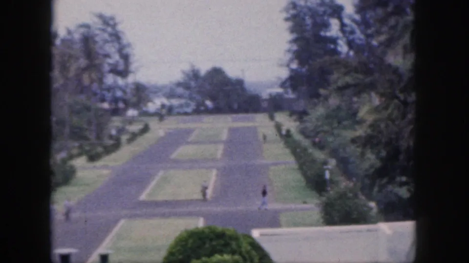 Soft 8mm view from Hawaii in 1959 showing intersecting paved paths, clipped lawns, tropical foliage, distant cars, and several small figures.