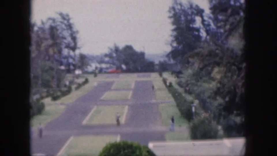 1959 Hawaii frame with a broad paved walkway system, trimmed lawns, tall trees, a low wall, distant cars, and small pedestrians.