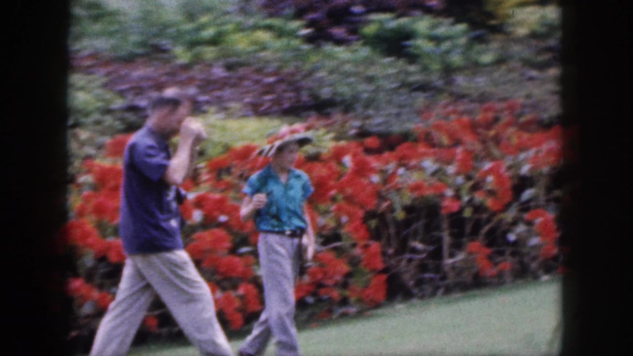 Man with camera walks beside a woman in a hat along a garden path with red flowers.