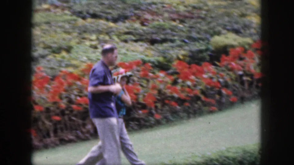The man carrying a camera passes close to frame as the woman is partly hidden by flowers in the Hawaii garden scene.