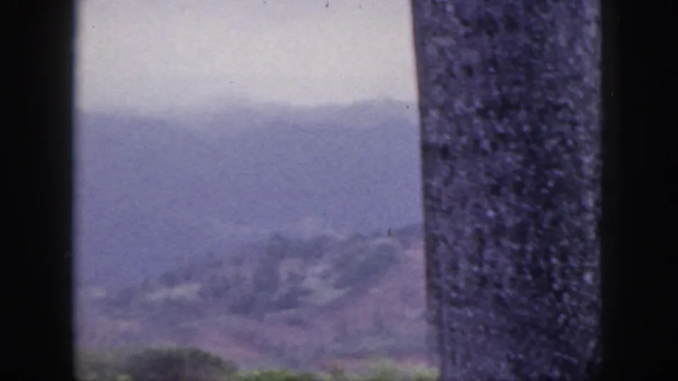 1959 Hawaii 8mm frame showing hazy mountain ridges through a dark window frame with a textured pillar in the foreground.
