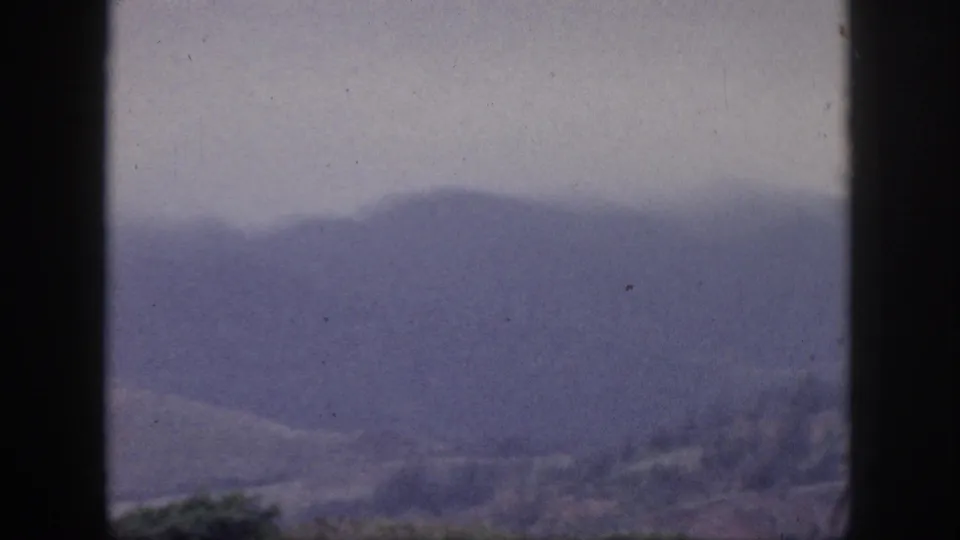 1959 Hawaii home-movie frame with distant purple-tinged mountains seen past dark vertical window bars.