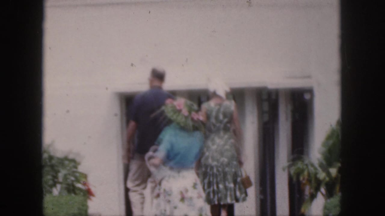 Adults in dresses and hats walk toward glass doors on a white building.