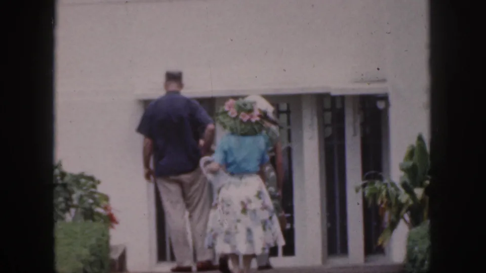 1959 Hawaii frame of adults seen from behind, including a woman carrying pink flowers near a white building.