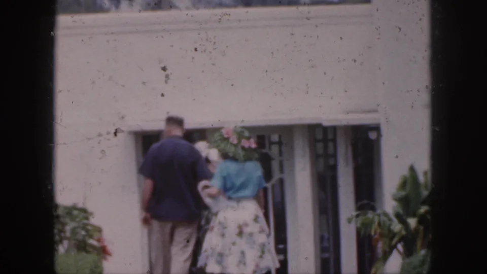 Adults in mid-century clothing approach a white building entrance, with tropical plants framing the walkway.