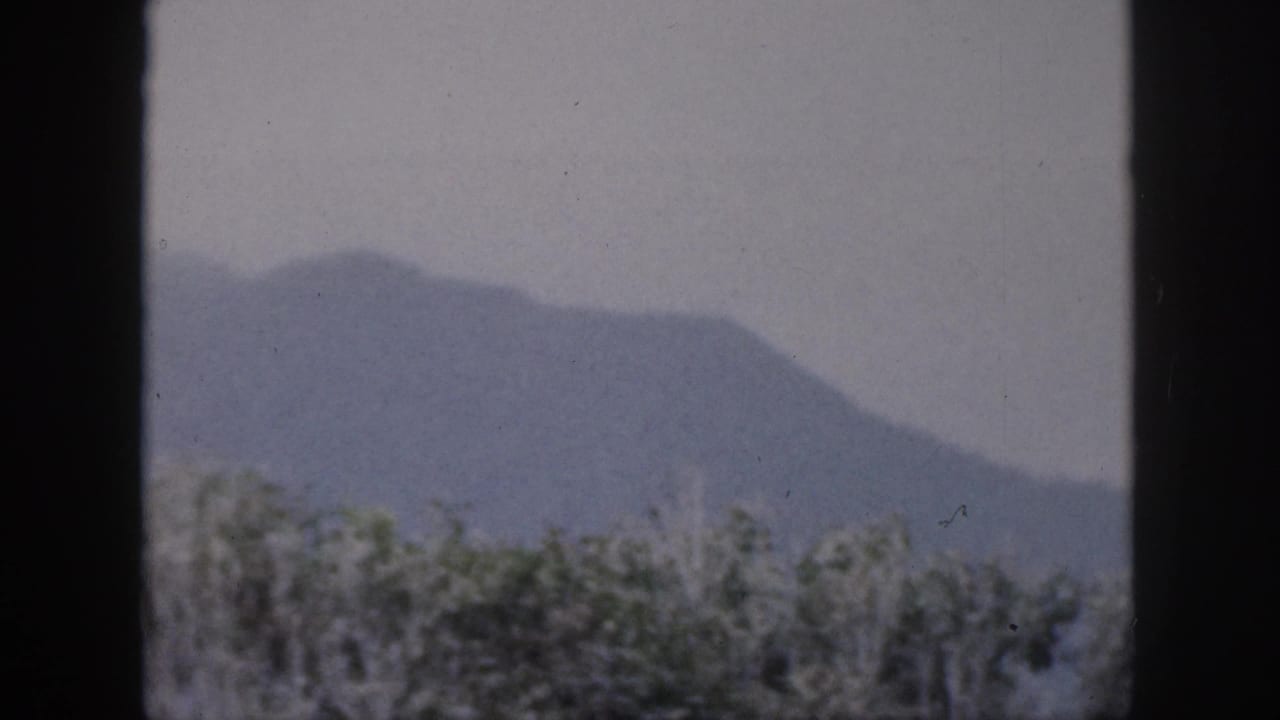 1959 Hawaii 8mm frame showing a soft blue-gray mountain ridge beyond tree tops, divided by a dark window or doorway frame.