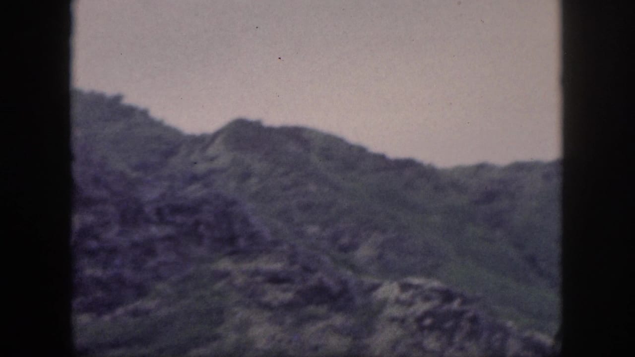 Vintage 8mm Hawaii frame showing rocky green mountain ridges through a dark window edge under an overcast sky.