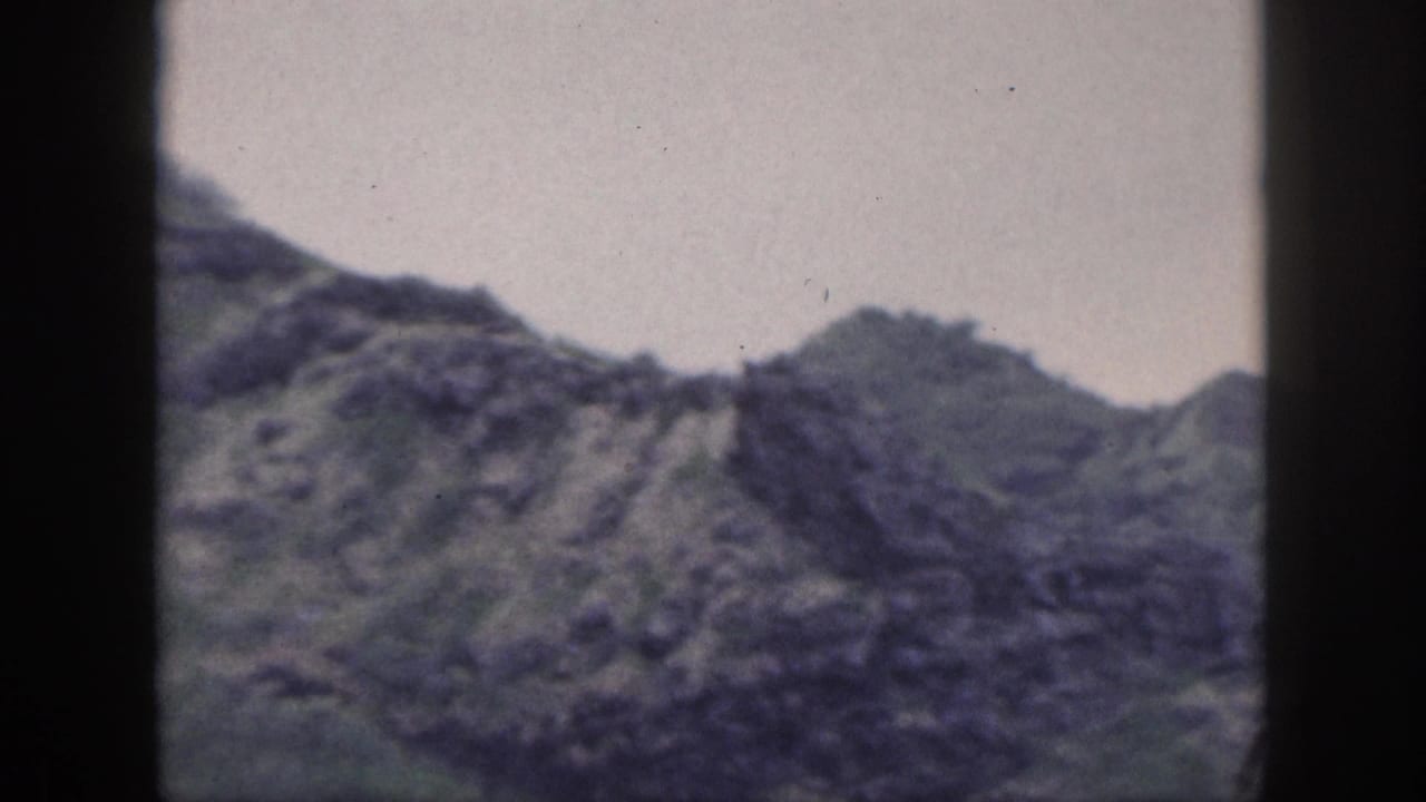 Archival Hawaii frame with a jagged mountain ridge, muted green slopes, pale sky, and visible film gate shadows.