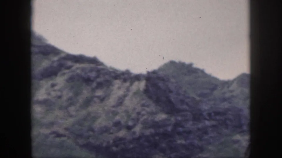 Archival Hawaii frame with a jagged mountain ridge, muted green slopes, pale sky, and visible film gate shadows.
