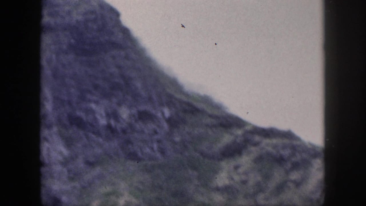Soft 8mm frame from Hawaii showing a steep, dark rocky slope with sparse green areas and an overcast sky.