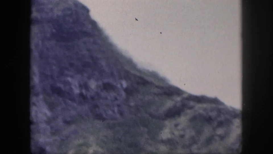 Soft 8mm frame from Hawaii showing a steep, dark rocky slope with sparse green areas and an overcast sky.