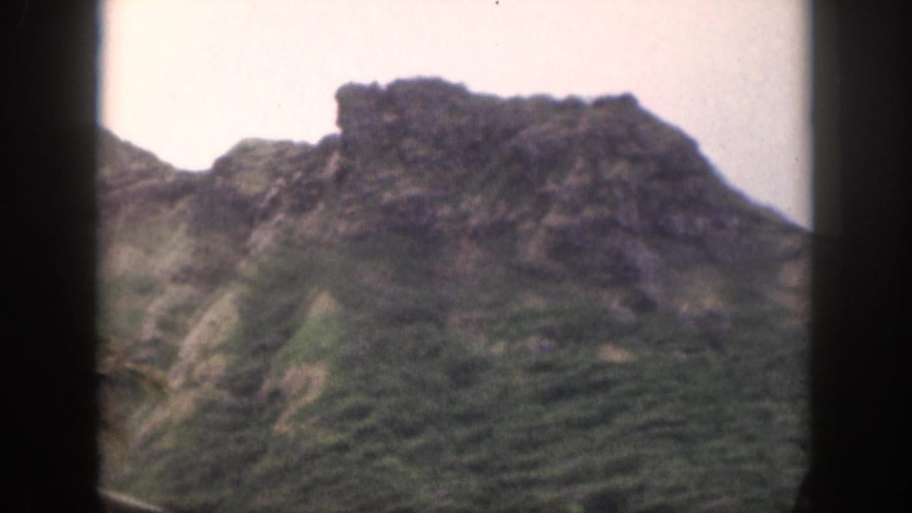 1959 Hawaii 8mm frame showing a steep vegetated slope and dark rocky ridge, with soft focus and faded color.