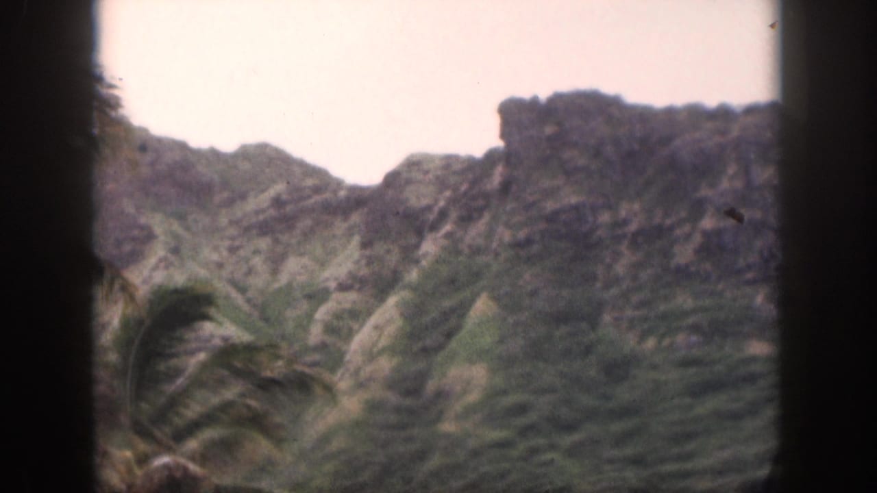 1959 Hawaii 8mm frame with palm fronds in front of rugged green mountain ridges and rocky outcrops.
