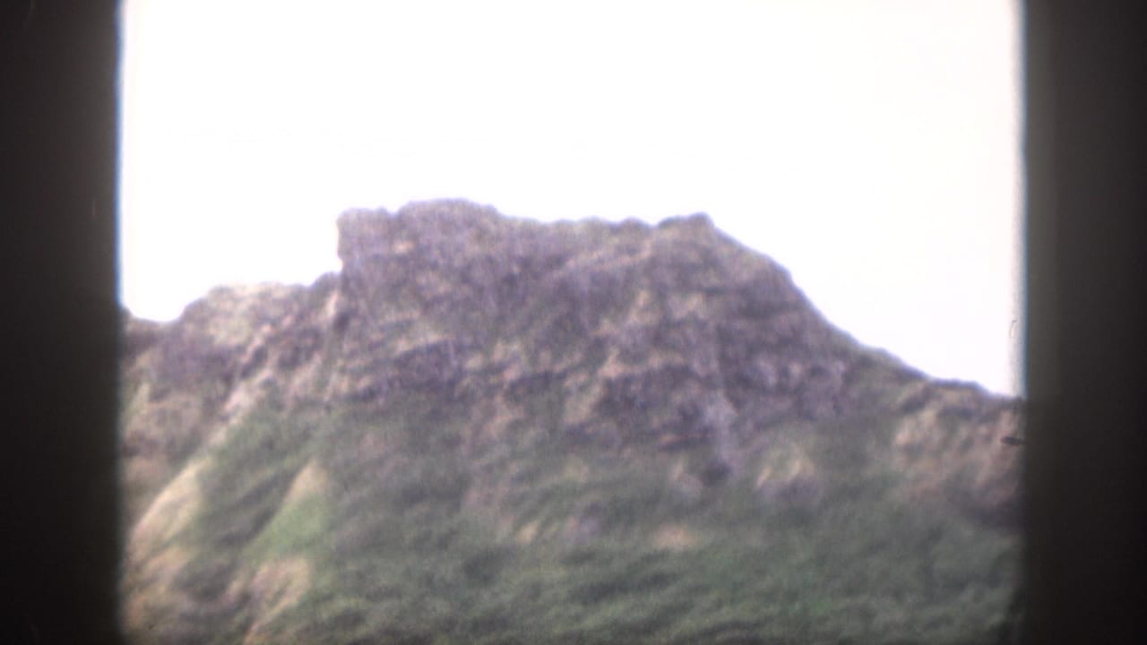 1959 Hawaii home-movie frame focused on a rough mountain ridge and green slopes, with bright sky and dark side borders.