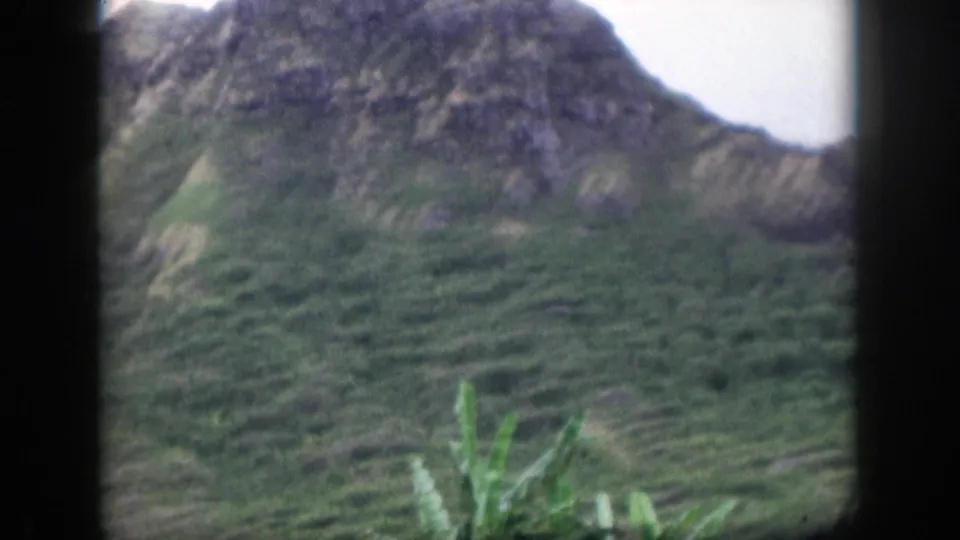 1959 Hawaii travel frame with a steep green mountain slope, rocky upper ridge, and tropical plants in the foreground.