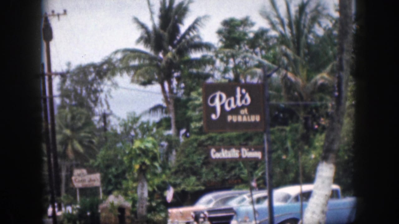 1959 Hawaii 8mm frame showing Pat's at Punaluu signage, parked mid-century cars, palm trees, utility poles, and a hazy mountain backdrop.