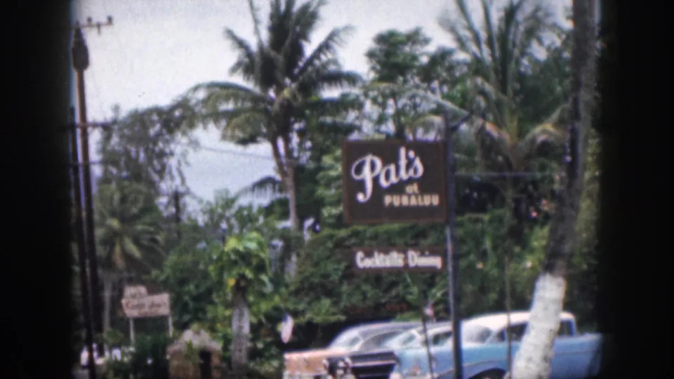 1959 Hawaii 8mm frame showing Pat's at Punaluu signage, parked mid-century cars, palm trees, utility poles, and a hazy mountain backdrop.