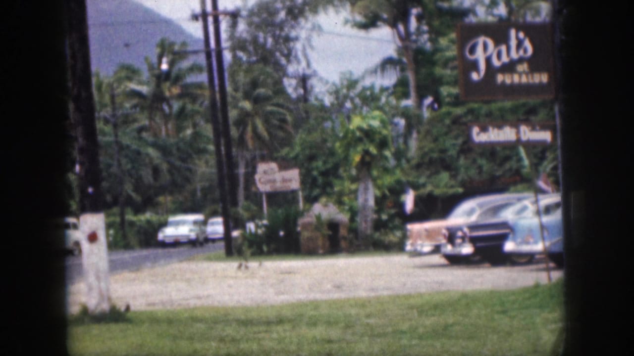 1959 Hawaii home-movie frame showing a grass foreground, roadside parking, colorful cars, Pat's at Punaluu signage, palms, and a pale mountain behind.