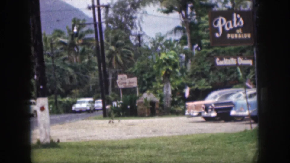 1959 Hawaii home-movie frame showing a grass foreground, roadside parking, colorful cars, Pat's at Punaluu signage, palms, and a pale mountain behind.