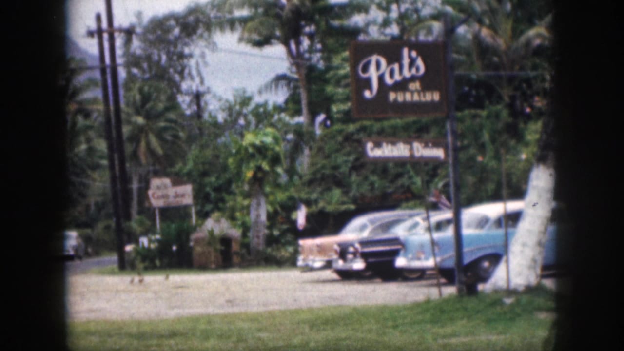 1959 Hawaii color 8mm view of Pat's at Punaluu signage above parked mid-century cars, with palms, grass, road edge, and tropical foliage.