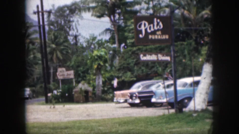 1959 Hawaii color 8mm view of Pat's at Punaluu signage above parked mid-century cars, with palms, grass, road edge, and tropical foliage.