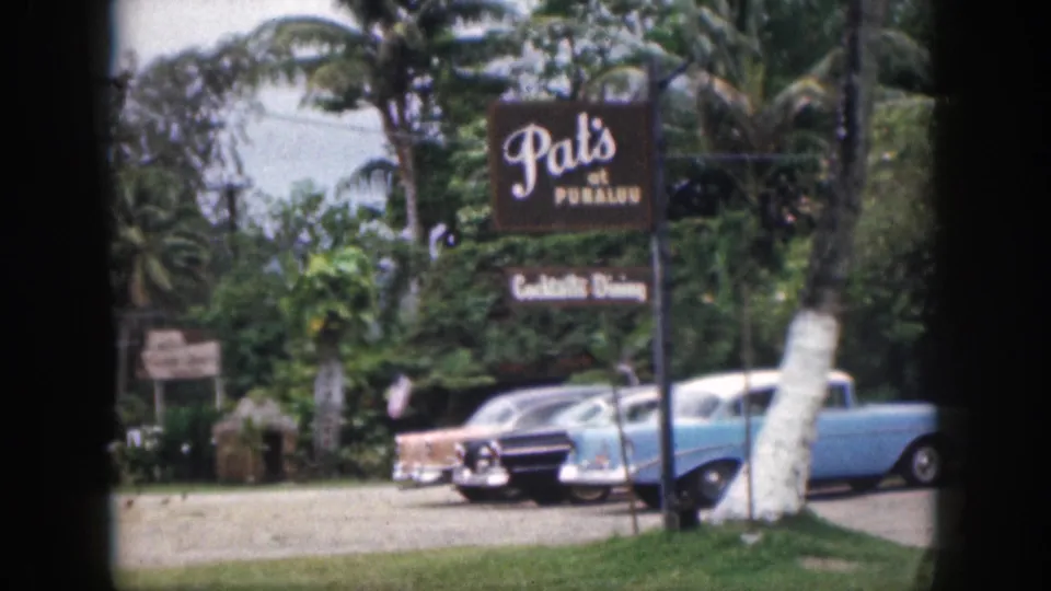 1959 Hawaii home-movie frame showing Pat's at Punalu'u signage, parked classic cars, palms, and roadside greenery.