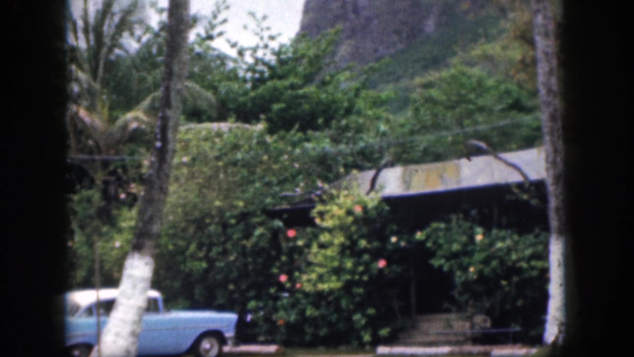 1959 Hawaii home-movie view of a pale blue car near dense tropical shrubs, palms, and a low building at the base of mountains.