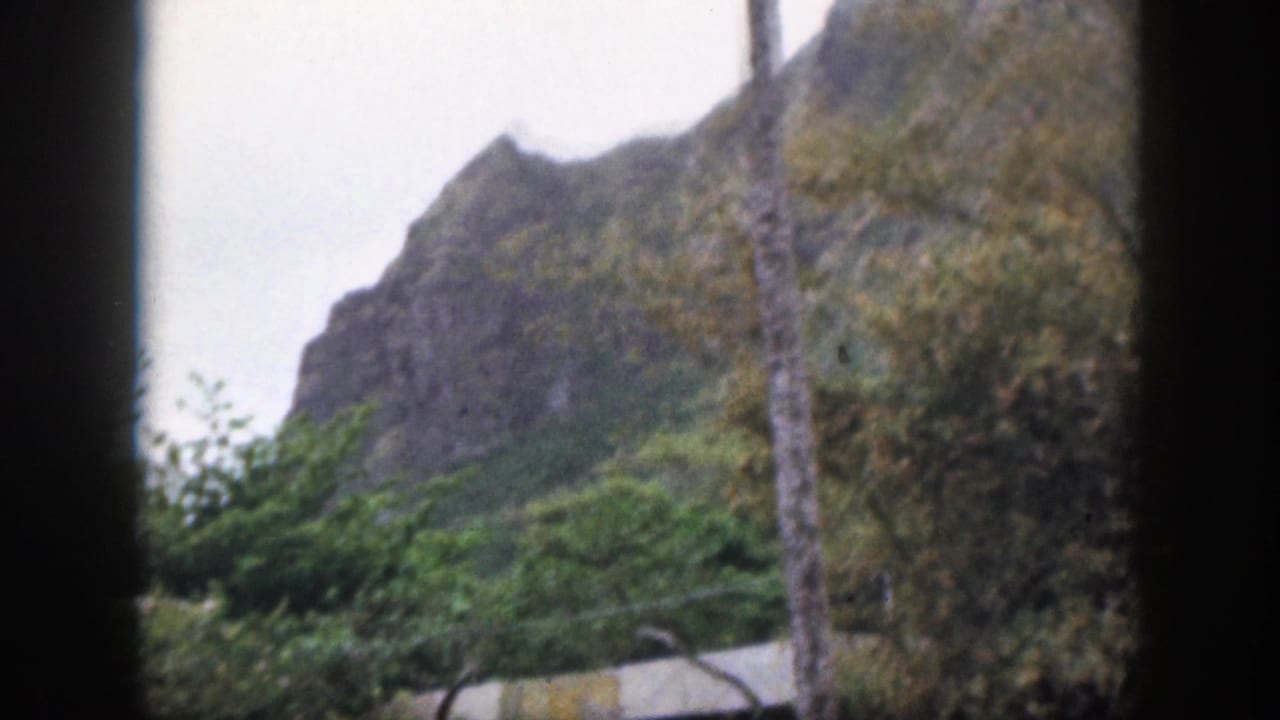 1959 Hawaii 8mm frame showing a green rocky mountain, trees and a pale overcast sky behind dark foreground edges.