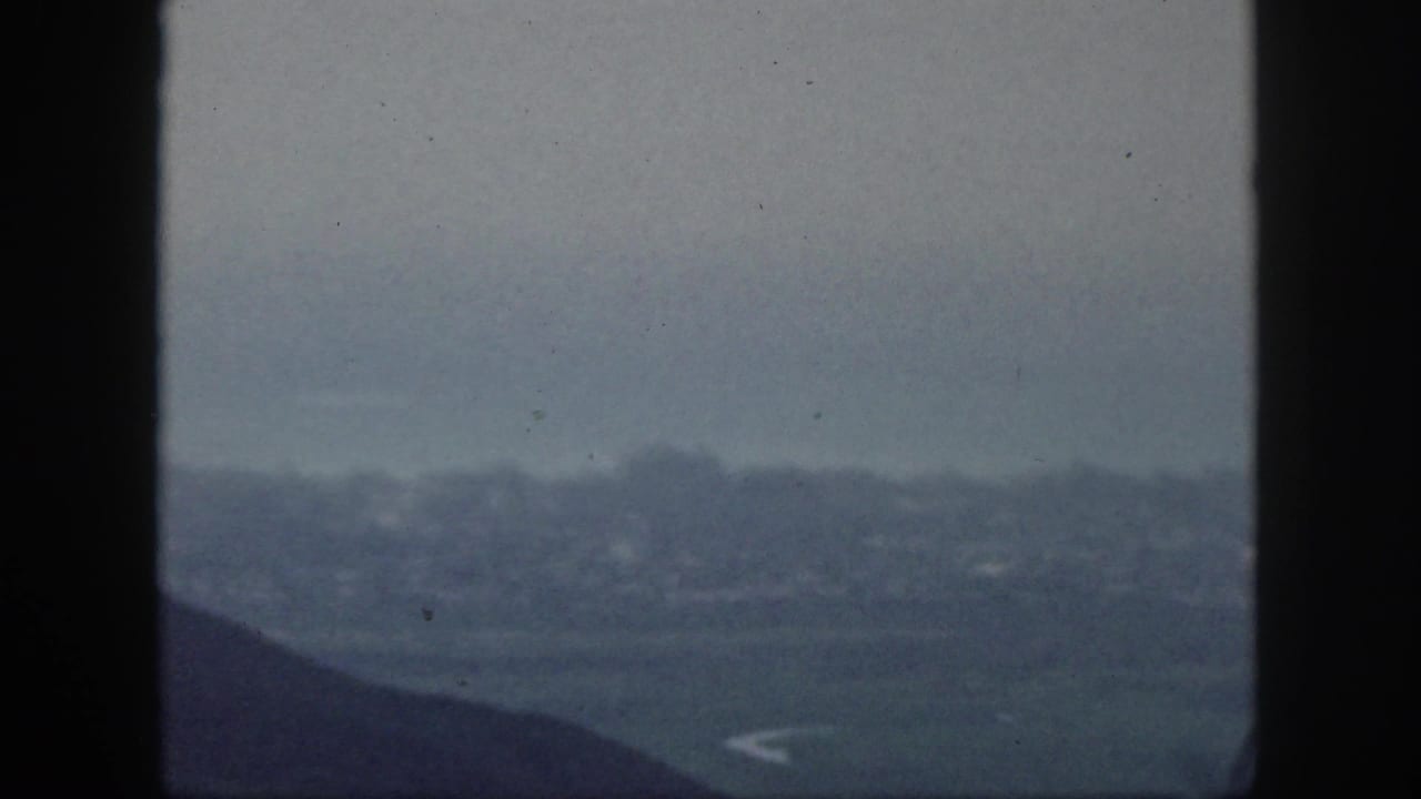 1959 Hawaii 8mm overlook toward a hazy distant settlement or shoreline beneath dark vertical framing.