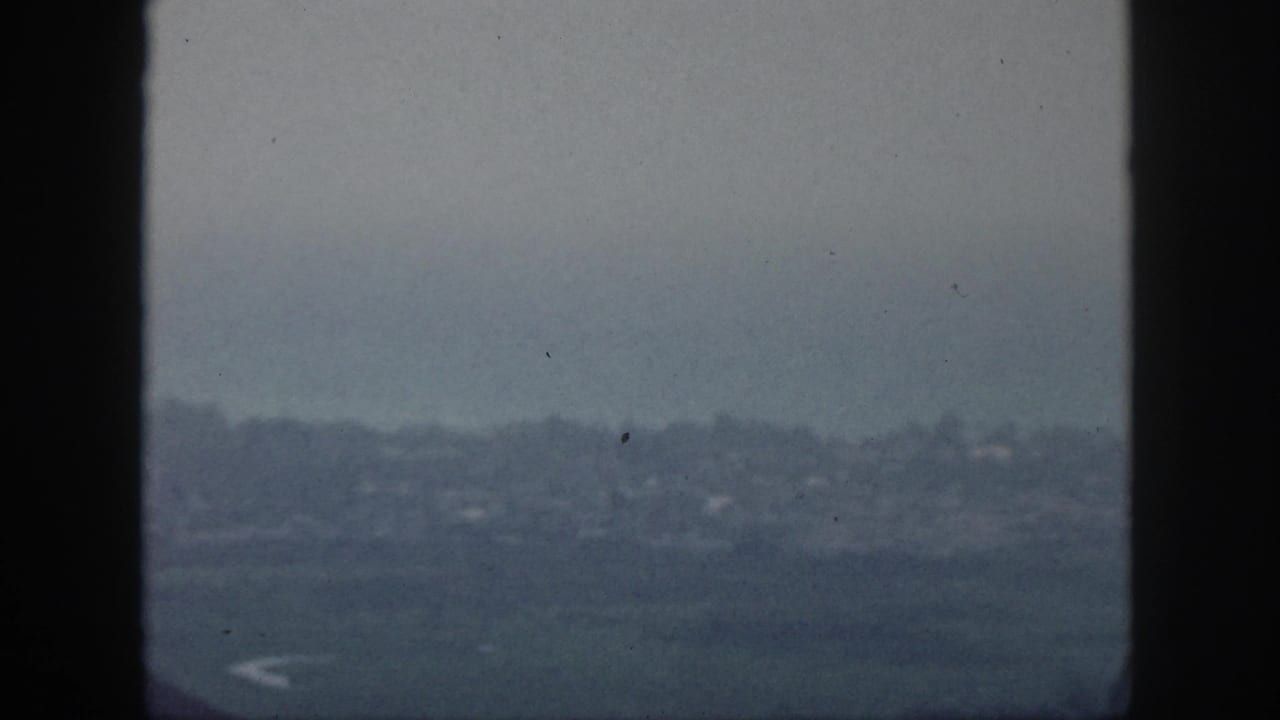 A muted 1959 Hawaii home-movie view looks past window framing toward a hazy shoreline and faint buildings.