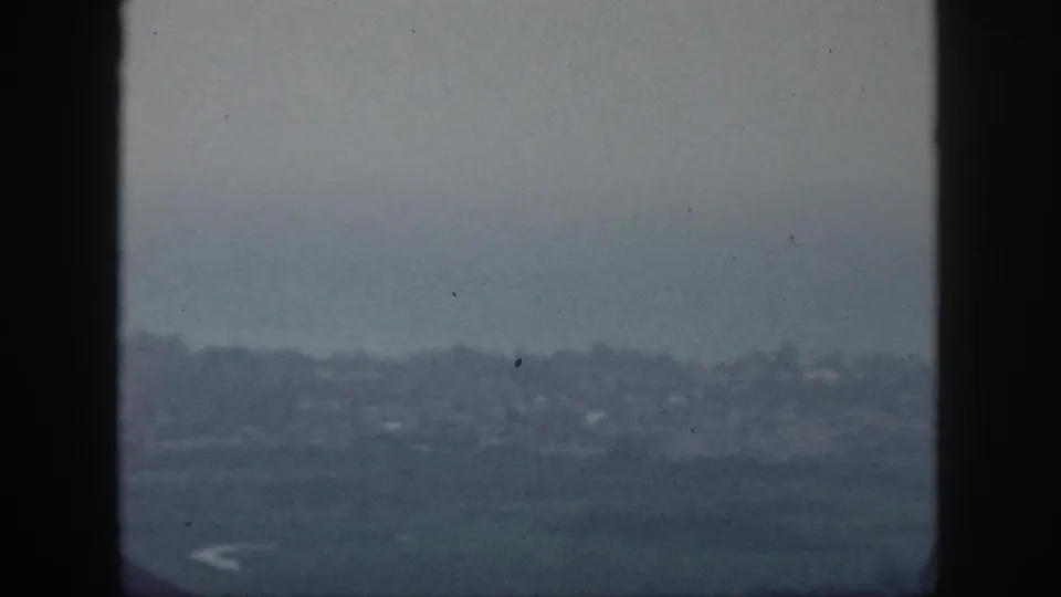 A muted 1959 Hawaii home-movie view looks past window framing toward a hazy shoreline and faint buildings.