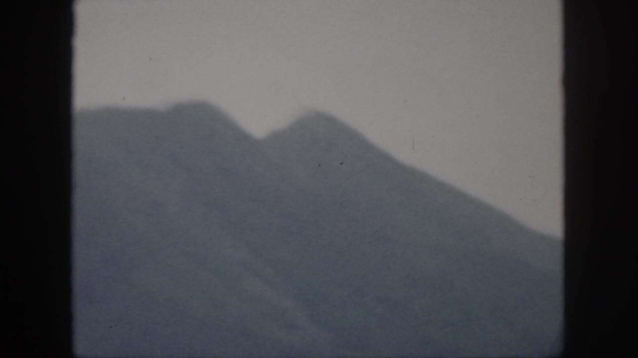 1959 Oahu home-movie frame showing distant rugged mountain peaks beneath a hazy, pale sky, bordered by the 8mm film scan.