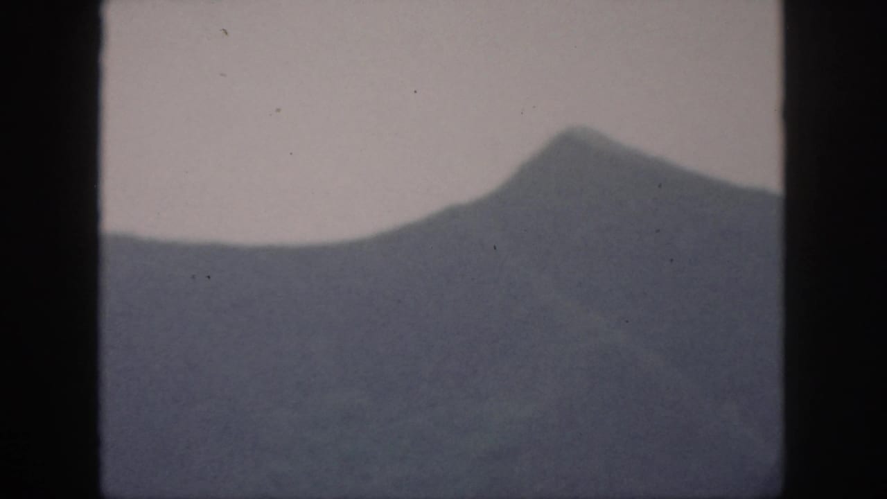1959 Oahu home-movie frame showing a steep mountain slope and central peak below a pale, overcast sky.