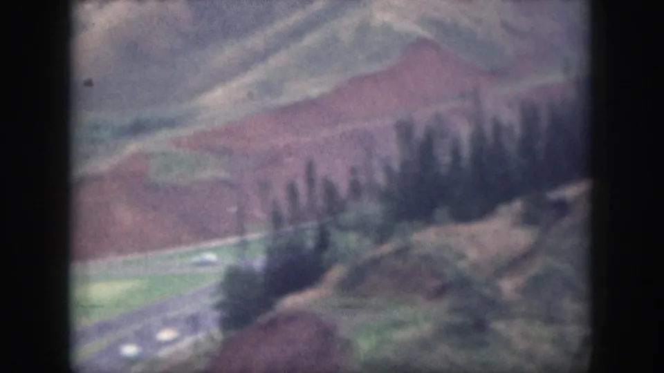 1959 Oahu 8mm frame showing a road curving through rolling hills, reddish earth, green patches, and small cars below.